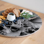 A round serving board with a black-and-white floral peony print, styled with bread, crackers, blueberries, a small white pitcher, cheese, and rosemary on a light wood table.
