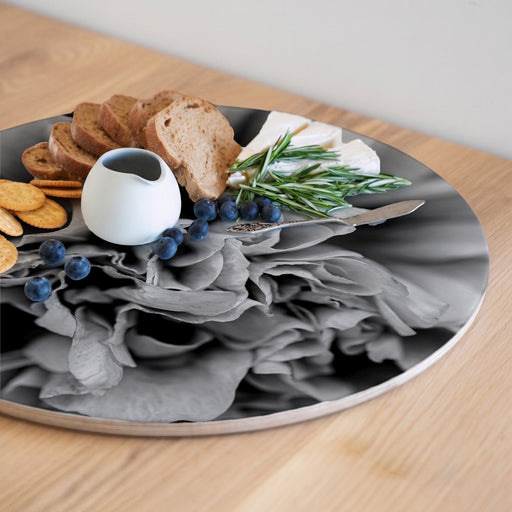 A round serving board with a black-and-white floral peony print, styled with bread, crackers, blueberries, a small white pitcher, cheese, and rosemary on a light wood table.