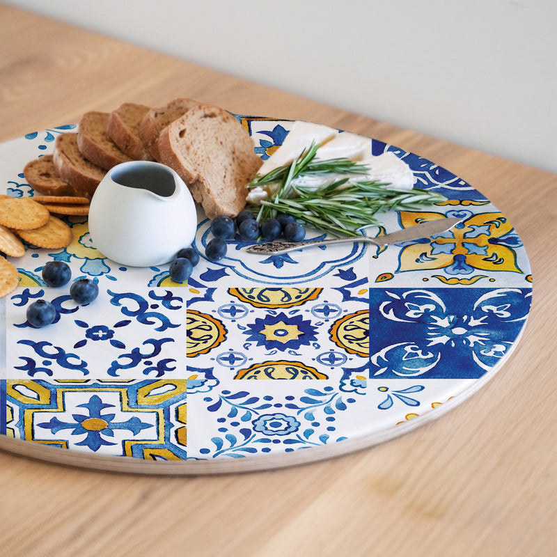 A round wooden serving board featuring a blue-and-yellow watercolour Lisbon tile print, styled with bread, blueberries, crackers, and a small white pitcher on a light wood table.