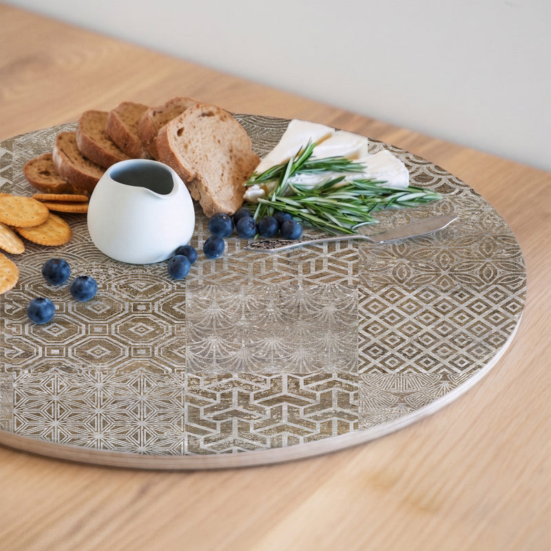 A wooden serving board featuring a brown modern tile pattern print, styled with crackers, bread, blueberries, cheese, rosemary, and a small white pitcher on a light wood table.