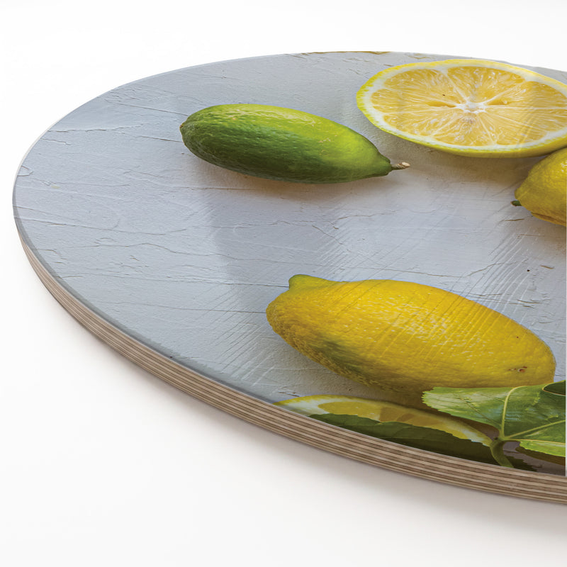 A round wooden serving board with a printed or painted citrus-themed design, featuring yellow lemons, a green lime, and green leaves on a light gray surface, photographed on a white background.