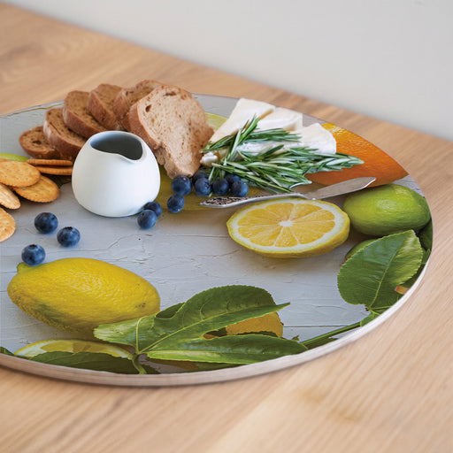 A round wooden serving board featuring a yellow-and-orange citrus-on-white print, styled with bread, crackers, blueberries, a small white pitcher, cheese, rosemary, and sliced lemons on a wooden table.