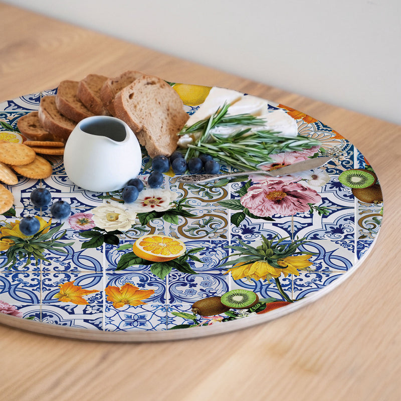 A round wooden serving board featuring a blue Lisbon tile with a lemons-and-flowers print, decorated with illustrations of fruit, flowers, and leaves. The platter is styled with food items including bread, crackers, blueberries, a small white pitcher, and a knife on a light wooden table.