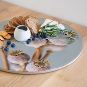 A round wooden serving board featuring a purple giant poppy seed bunch print styled with crackers, blueberries, bread, cheese, rosemary, and a small white pitcher on a light wood tabletop.