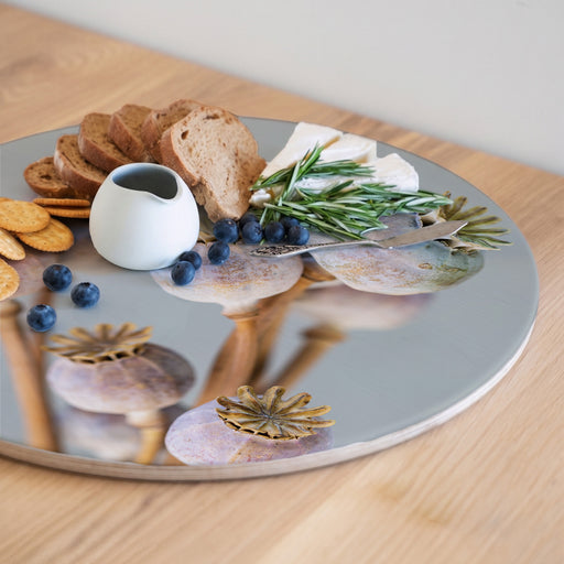 A round wooden serving board featuring a purple giant poppy seed bunch print styled with crackers, blueberries, bread, cheese, rosemary, and a small white pitcher on a light wood tabletop.