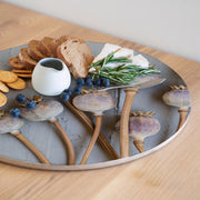 A round wooden serving board featuring a purple giant poppy seed print, styled with bread, crackers, blueberries, cheese, rosemary, and a small white pitcher on a light wood table.