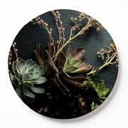 Round serving board featuring a printed botanical arrangement of succulents and dried stems on a dark textured background, shown against a clean white wall.
