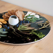 A round printed serving board printed with a green aloe and leaves pattern, styled with crackers, bread, blueberries, cheese, rosemary, and a small white ceramic pitcher on a light wood table.