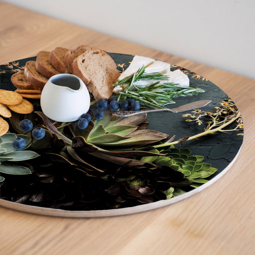 A round printed serving board printed with a green aloe and leaves pattern, styled with crackers, bread, blueberries, cheese, rosemary, and a small white ceramic pitcher on a light wood table.