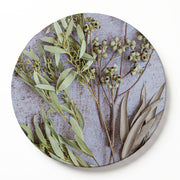 A round wooden serving board featuring a botanical print of eucalyptus leaves and green amber nuts on a textured light grey background, shown against a clean white backdrop.