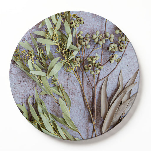 A round wooden serving board featuring a botanical print of eucalyptus leaves and green amber nuts on a textured light grey background, shown against a clean white backdrop.