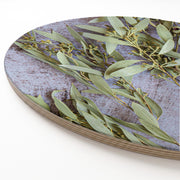 A round wooden serving board with a printed botanical design of green eucalyptus leaves and amber nuts on a distressed grey-blue background, shown on a white surface.