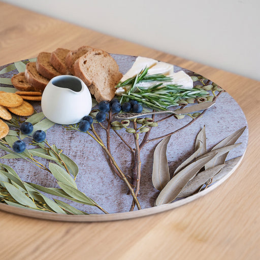 A round wooden serving board with a botanical print, styled with crackers, bread, cheese, blueberries, rosemary, and a small white ceramic pitcher on a light wood table.