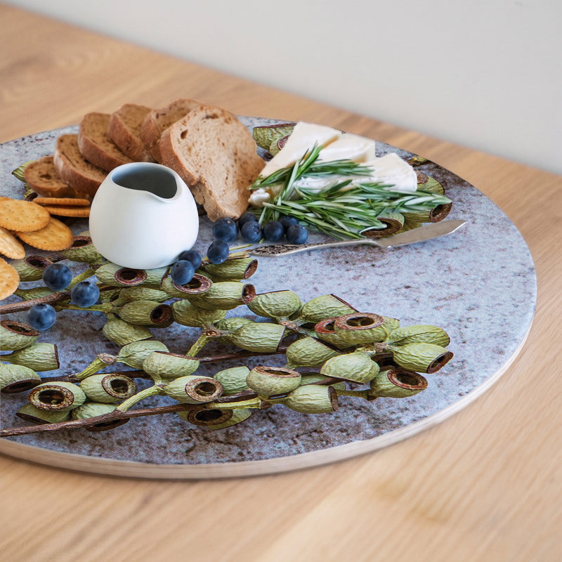 A round wooden serving board featuring a green natural ambernut print styled with bread, crackers, cheese, blueberries, rosemary, and green pods, set on a light wood table.