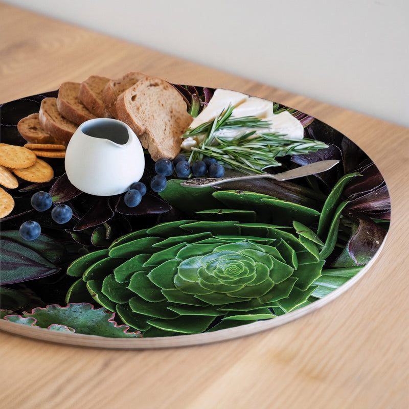 A round printed serving board printed with a succulent and leaf pattern, styled with sliced bread, crackers, blueberries, a small white pitcher, and rosemary on a wooden table.