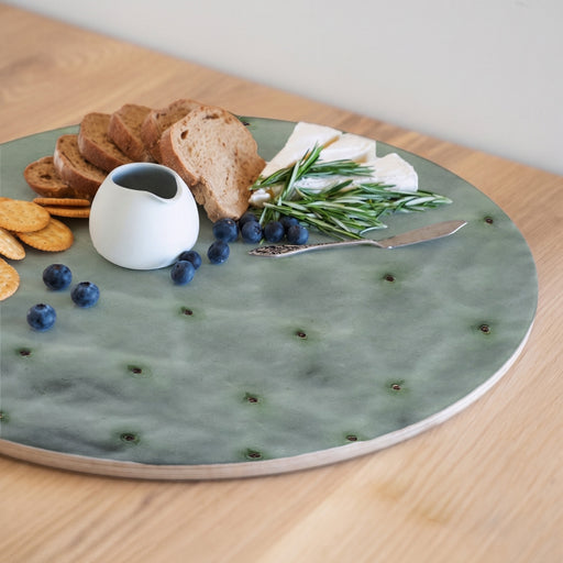 A round wooden serving board featuring a green prickly pear leaf print, styled with bread, crackers, blueberries, cheese, rosemary, and a small white pitcher on a light wood table.