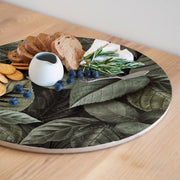 A round wooden serving board featuring an all-over monochromatic green botanical leaf print, styled with bread, crackers, blueberries, a small white pitcher, cheese, and rosemary on a light wood table.
