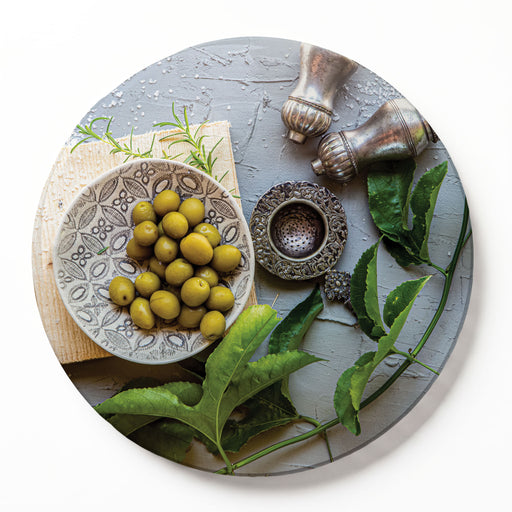 A round wooden serving board featuring a silver salt shaker and a green olive plate print, photographed against a white background.