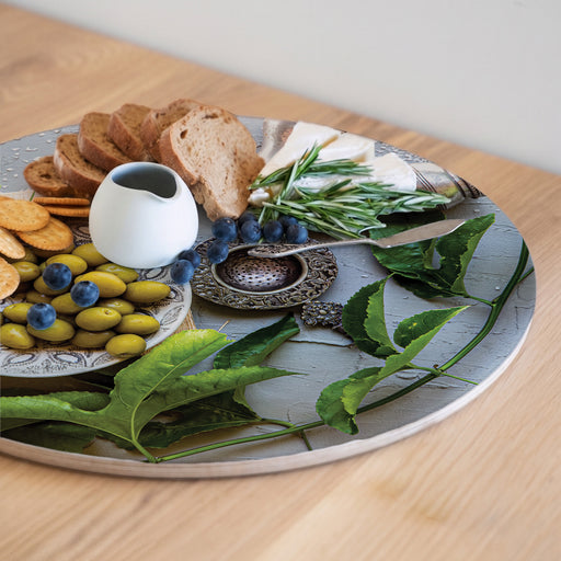 A round wooden serving board featuring a silver salt shaker and a green olive plate print styled with food items including sliced bread, olives, blueberries, a small white pitcher, a metal dish, and green leaves on a light wood tabletop.