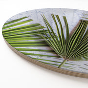 Round print serving board featuring a close-up photograph of green palm leaves over a light grey marbled background, shown at an angle on a white surface.