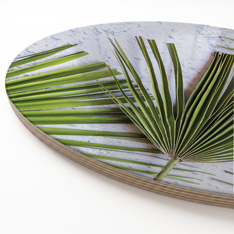 Round print serving board featuring a close-up photograph of green palm leaves over a light grey marbled background, shown at an angle on a white surface.
