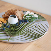 A round printed serving board with a light grey and green palm leaf pattern, styled with bread, crackers, blueberries, cheese, rosemary, and a small white pitcher on a wooden table.