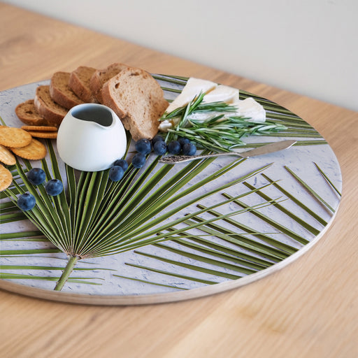 A round printed serving board with a light grey and green palm leaf pattern, styled with bread, crackers, blueberries, cheese, rosemary, and a small white pitcher on a wooden table.
