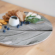 A round wooden serving board featuring a grey monochrome leaf print, styled with bread, cheese, blueberries, crackers, rosemary, and a small white creamer on a light wood table.