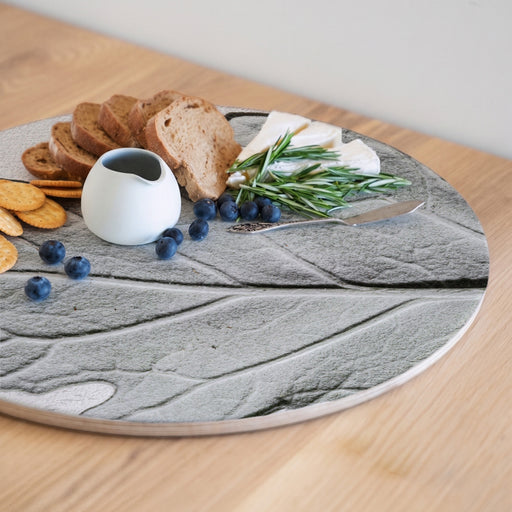 A round wooden serving board featuring a grey monochrome leaf print, styled with bread, cheese, blueberries, crackers, rosemary, and a small white creamer on a light wood table.