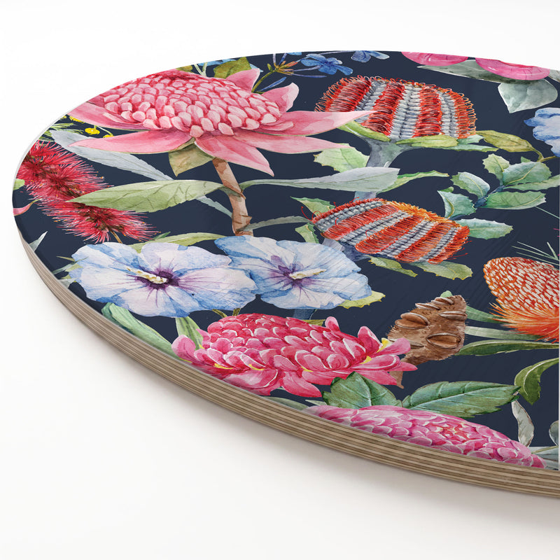 A round wooden serving board featuring a vibrant floral print with pink, red, and blue flowers on a dark navy background, photographed on a clean white surface.