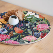 A round serving board with a colourful botanical print featuring pink flowers, green leaves, and blue berries on a dark navy blue background, styled with crackers, bread, cheese, rosemary, and a small white pitcher on a light wood table.