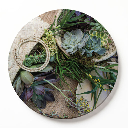 A round printed serving board featuring a botanical print of aloe, succulents, greenery, woven baskets, and a straw hat on a burlap background, photographed from above against a white backdrop.