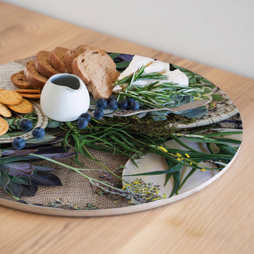 A round printed serving board with a botanical and burlap print, styled with bread, crackers, blueberries, a small white pitcher, cheese, and green herbs on a light wood table.