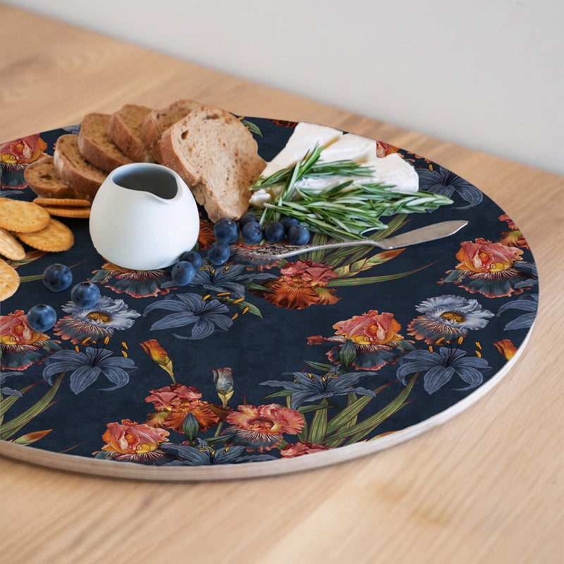A round printed floral serving board with a dark navy background and colourful flowers, styled with bread, crackers, blueberries, cheese, rosemary, and a small white pitcher on a light wood table.
