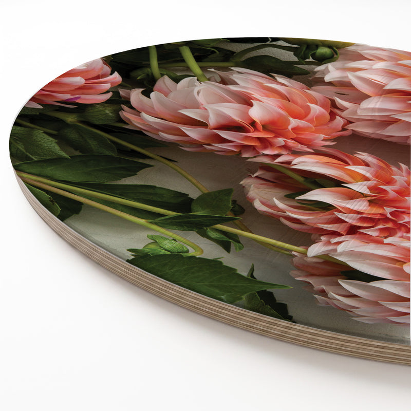 A round wooden serving board featuring a printed floral design of pink and orange dahlias with green leaves, shown on a clean white background.