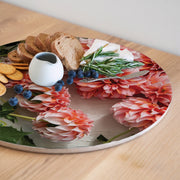 A round serving board with a floral print featuring large pink and orange dahlia flowers, styled with crackers, bread, blueberries, cheese, rosemary, and a small white pitcher on a light wood table.