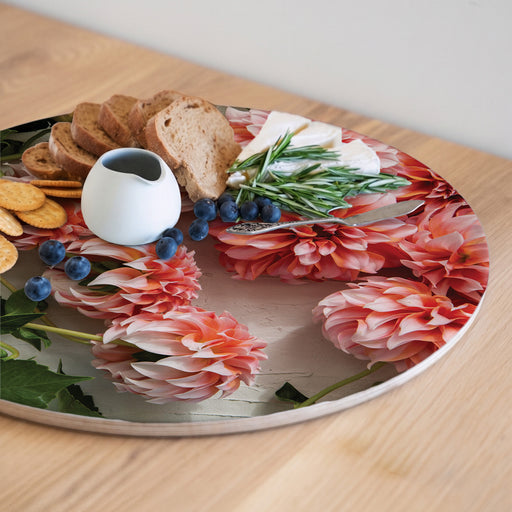 A round serving board with a floral print featuring large pink and orange dahlia flowers, styled with crackers, bread, blueberries, cheese, rosemary, and a small white pitcher on a light wood table.