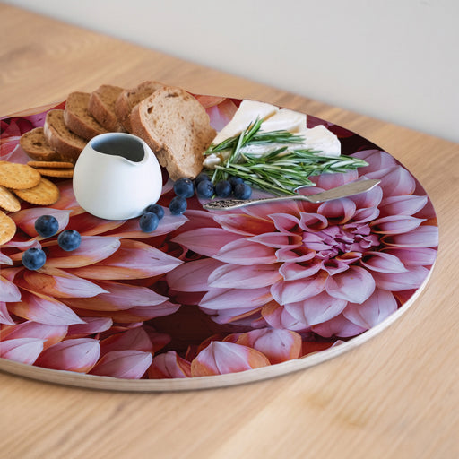 A round serving board with a large pink and orange dahlia floral print, styled with crackers, bread, blueberries, cheese, rosemary, and a small white pitcher on a light wood table.