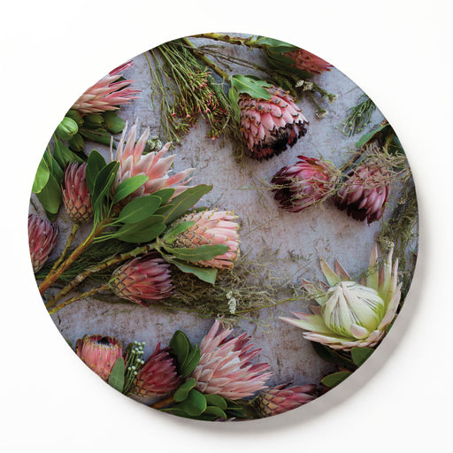 A round printed serving board featuring a botanical print of pink protea flowers and green leaves on a textured light grey background, shown against a clean white backdrop.