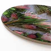 A round wooden serving board featuring a detailed photographic print of pink protea flowers and green leaves, shown at an angle on a white background.