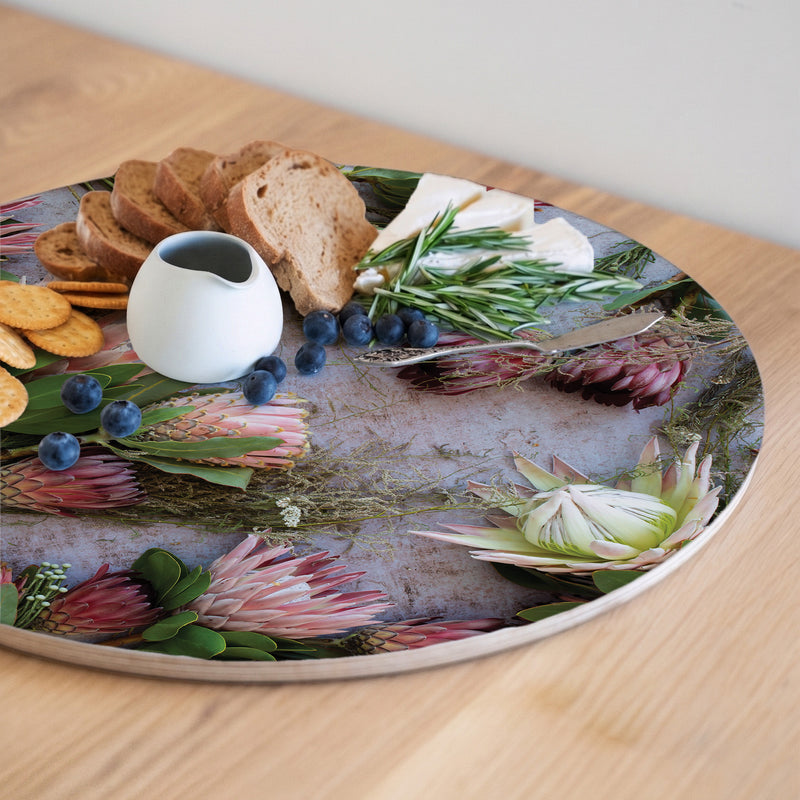 A round printed serving board with a botanical print of pink protea flowers and green leaves, styled with crackers, bread, blueberries, cheese, and a small white pitcher on a light wood table.