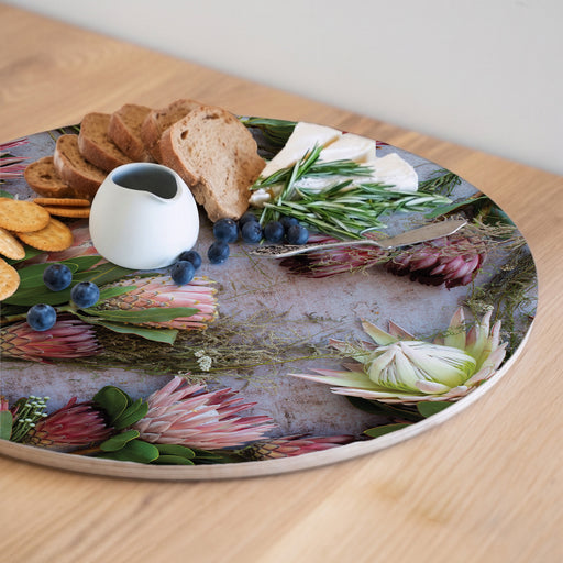 A round printed serving board with a botanical print of pink protea flowers and green leaves, styled with crackers, bread, blueberries, cheese, and a small white pitcher on a light wood table.