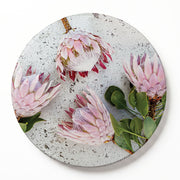 A round printed serving board featuring a photographic print of four pink protea flowers with green leaves on a textured light grey background, shown on a clean white surface.