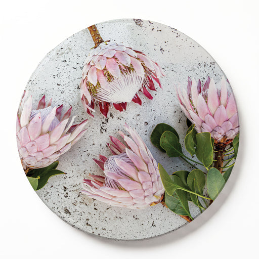 A round printed serving board featuring a photographic print of four pink protea flowers with green leaves on a textured light grey background, shown on a clean white surface.