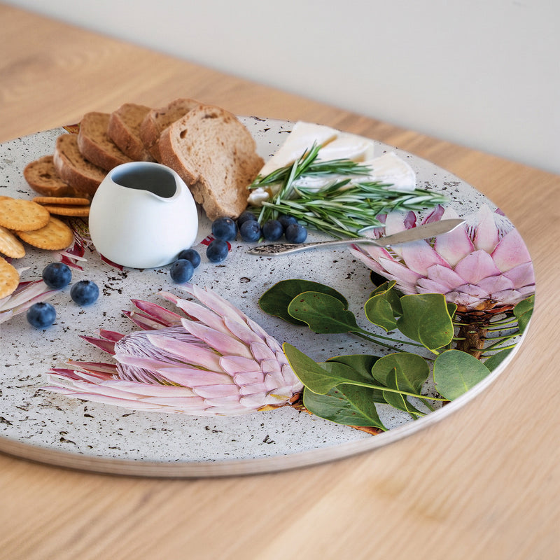 A round, pink protea on a light grey background, printed on a serving platter, styled with crackers, bread, blueberries, cheese, rosemary, and a small white pitcher set on a light wood table.