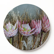 A round decorative serving board featuring a photographic print of pink protea flowers and dried grasses, shown against a clean white background.