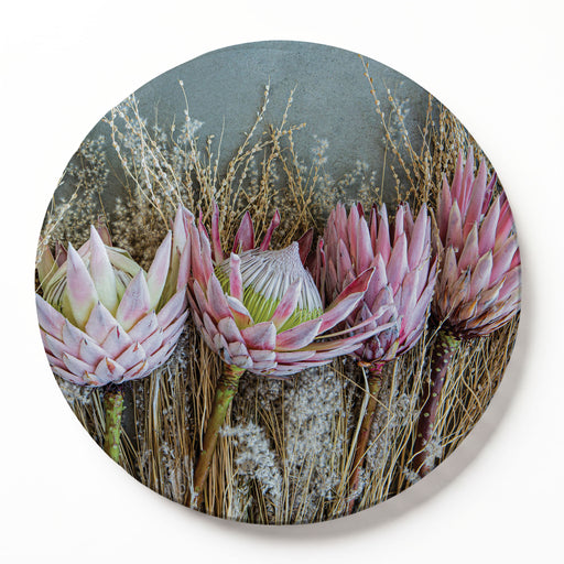 A round decorative serving board featuring a photographic print of pink protea flowers and dried grasses, shown against a clean white background.
