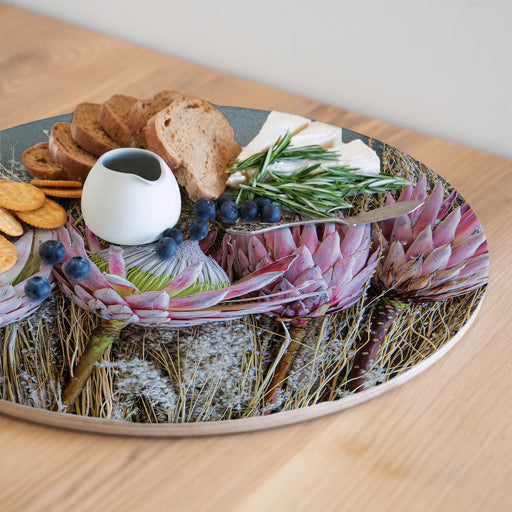 A round pink protea-printed serving platter filled with food items, including blueberries, bread, crackers, cheese, and rosemary, set on a light wood tabletop.