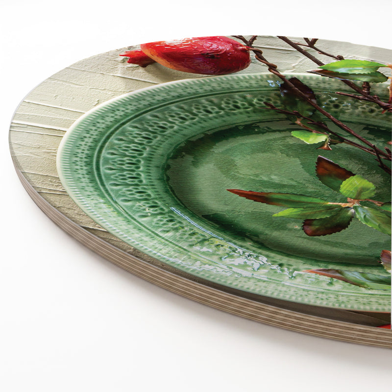 A close-up of a round wooden serving board featuring a red pomegranate on a green plate print.