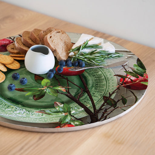 A round wooden serving board featuring a red pomegranate on a green plate print, styled with a small white pitcher, blueberries, crackers, and bread slices on a light wood table.